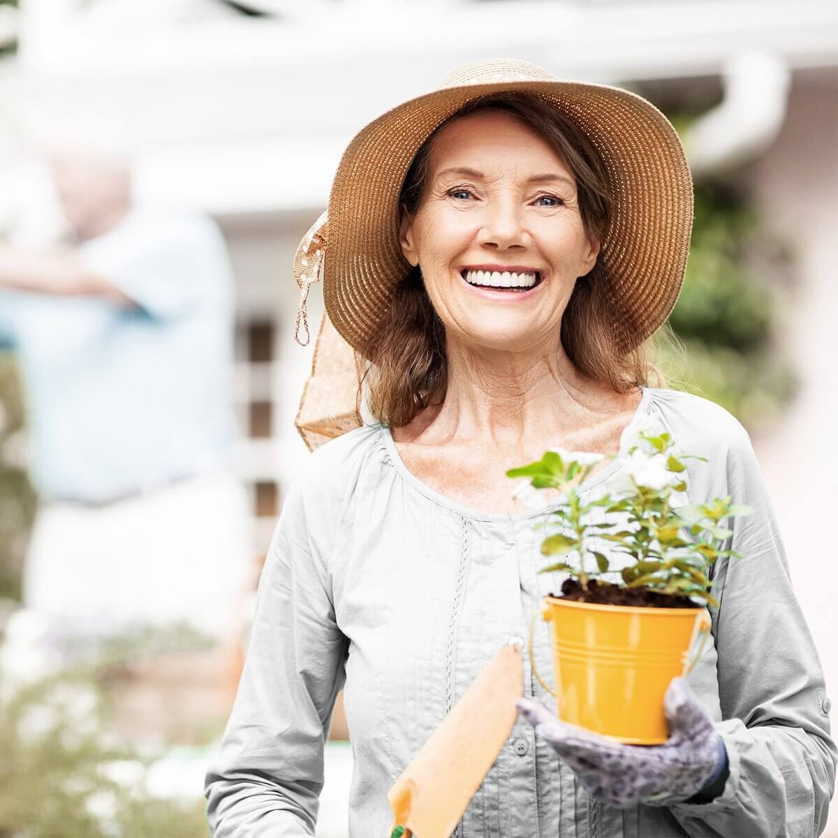 GELENK AKTIV forte: Frau steht lächelnd im Garten mit einer Topfpflanze und einer kleinen Schippe in den Händen