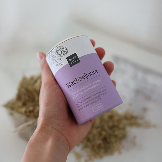 Eine weibliche Hand hält eine Dose mit Wechseljahrekapseln von Pure Ella. Im Hintergrund sind verschwommen einige Kräuterzutaten zu sehen.